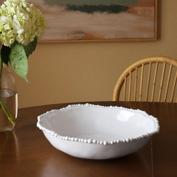 White decorative bowl on a wooden table with a vase of flowers and a chair in the background.