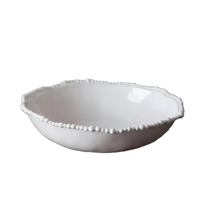 White decorative bowl on a wooden table with a vase of flowers and a chair in the background.