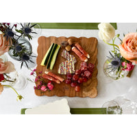 Acacia wood scalloped cutting board with charcuterie, grapes, and cucumber, surrounded by flowers.