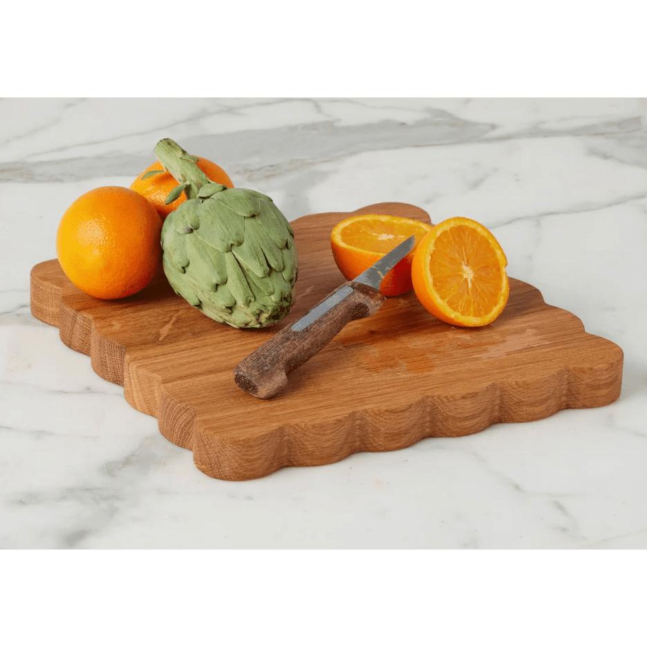 Acacia wood scalloped cutting board with oranges, artichoke, and knife on a marble surface.