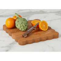 Acacia wood scalloped cutting board with oranges, artichoke, and knife on a marble surface.