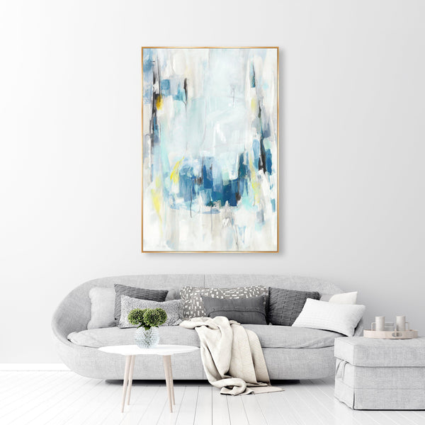 Abstract giclee canvas painting with blue, white, and yellow strokes in a gold frame above a gray sofa.