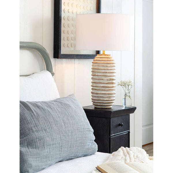 Distressed white-washed wood table lamp with carved ridges, a white drum shade, and brass finial on a nightstand.