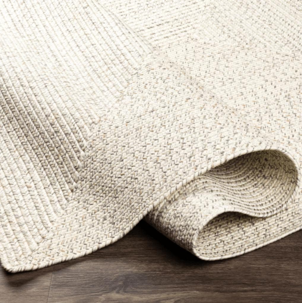 Chesapeake Bay Cream Outdoor Rug, braided polypropylene, partially rolled on a dark wood floor.