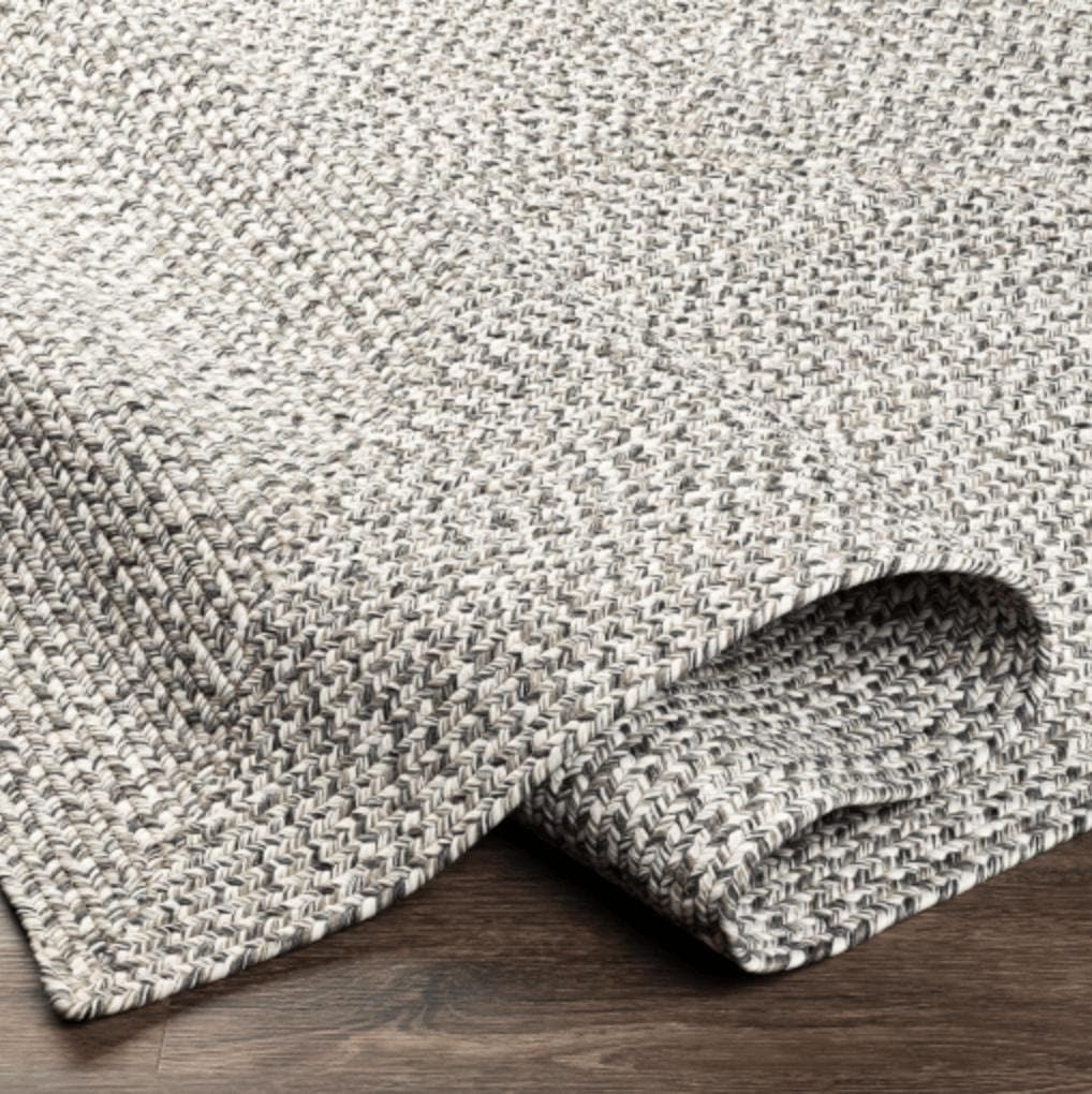 Gray braided polypropylene outdoor rug with a no-pile texture, partially rolled on a wood floor.