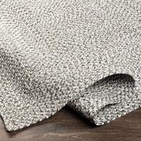 Gray braided polypropylene outdoor rug with a no-pile texture, partially rolled on a wood floor.
