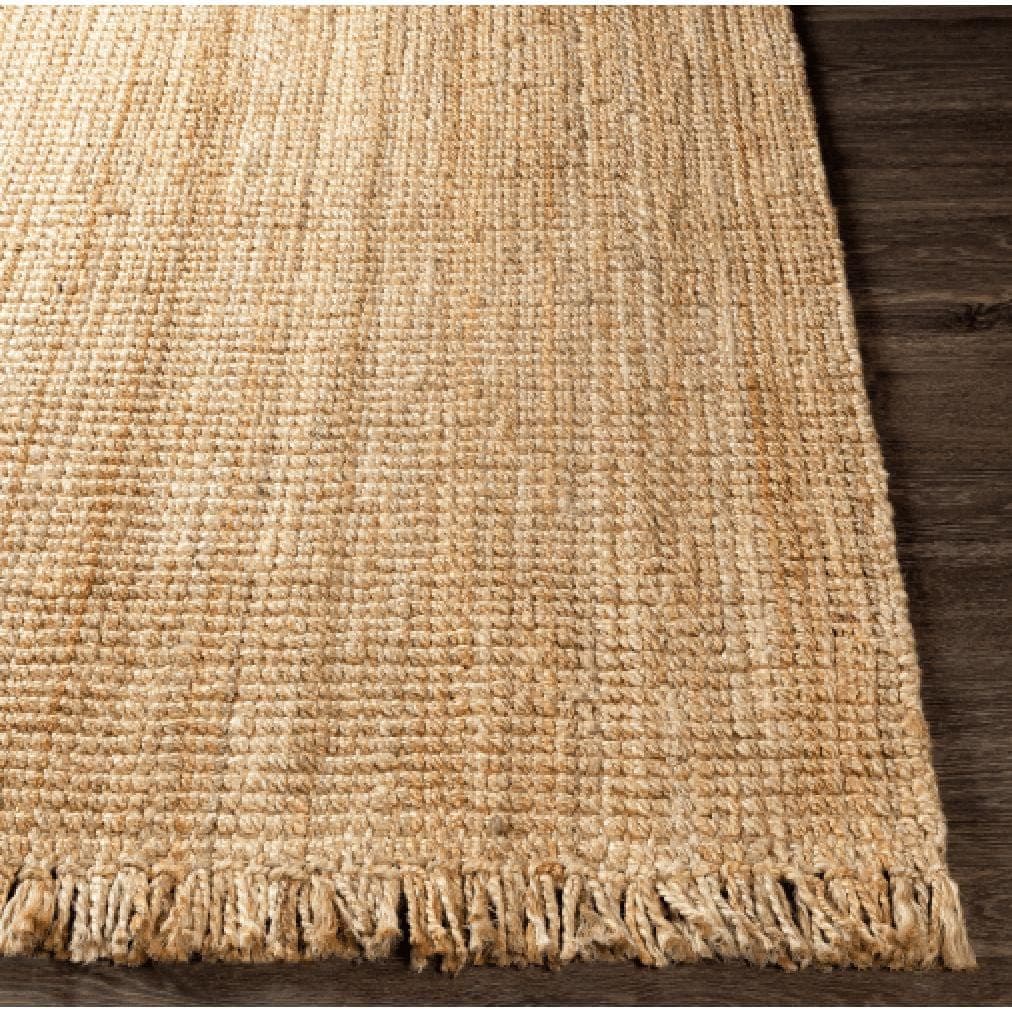 Light brown, hand-woven chunky jute rug with fringe, on a dark wood floor.