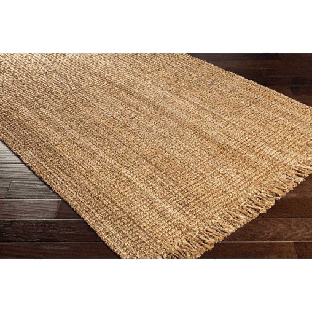Light brown, hand-woven chunky jute rug with fringe on a dark wood floor.