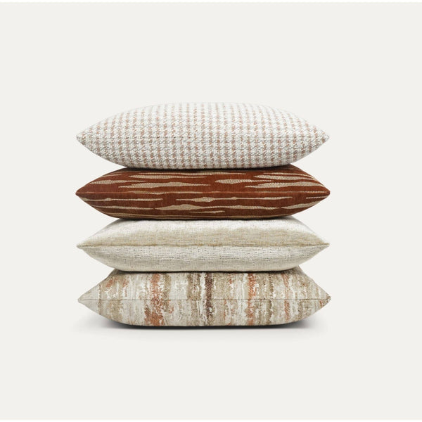 Stack of four luxury designer throw pillows in rust, cream, and beige with varied textures.