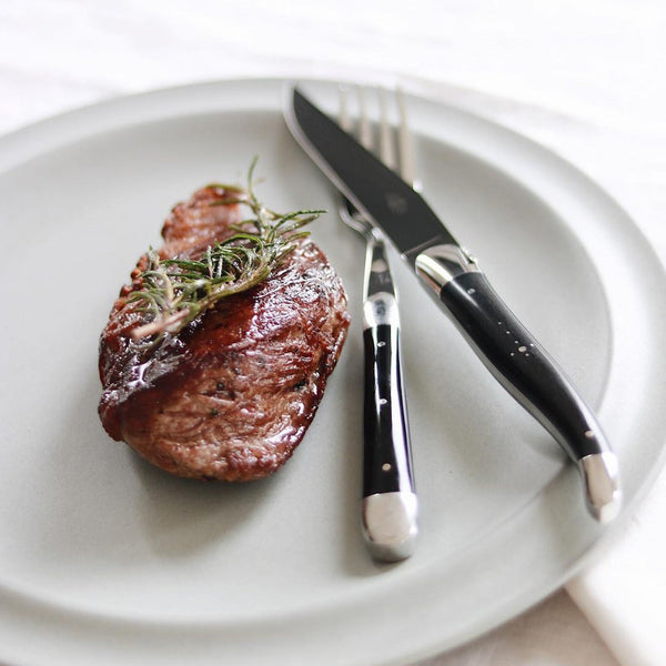 Steak with rosemary on a white plate with a knife and fork.