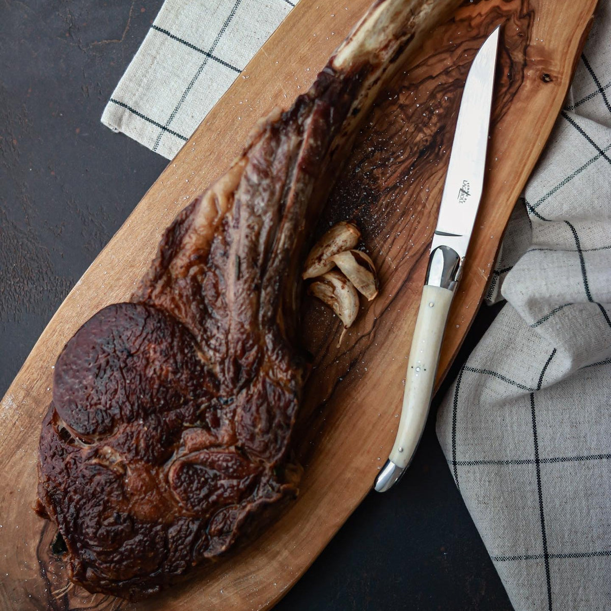 Grilled ribeye steak on a wooden cutting board with a knife, on a checkered cloth.