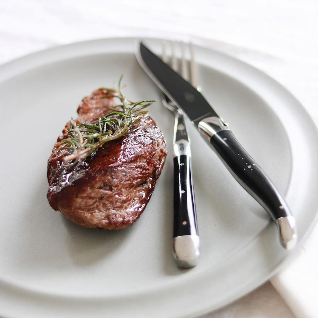 Steak with rosemary on a white plate with a knife and fork.