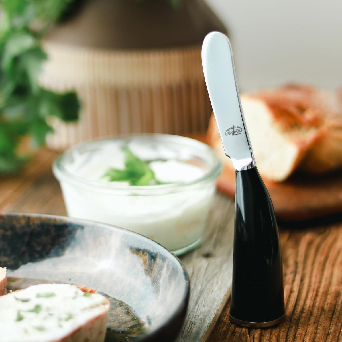 Butter knife with black handle on a wooden surface next to a jar of cream cheese and bread.
