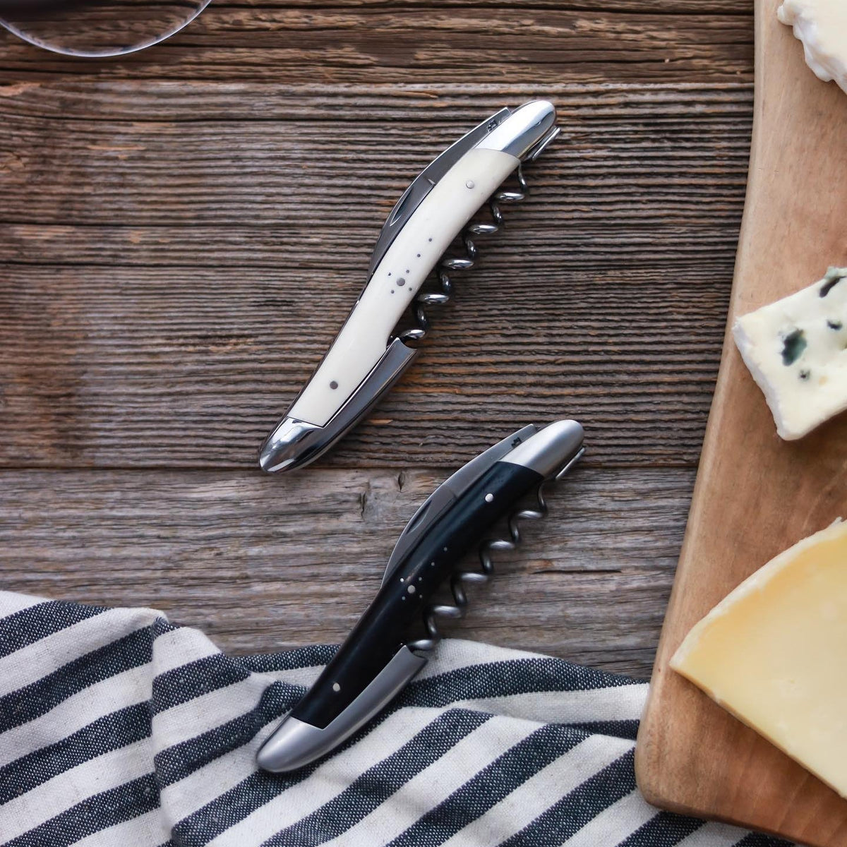 Two corkscrews on a wooden surface with a striped cloth and cheese in the background.