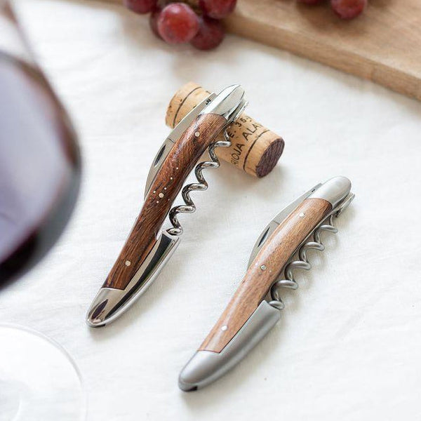 Two corkscrews with wooden handles on a white surface with grapes and a glass of red wine.