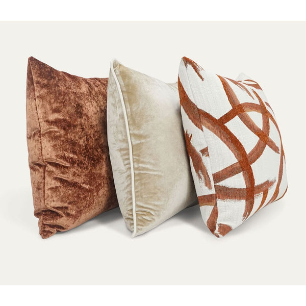 Three luxury throw pillows: a textured copper velvet, a cream velvet with piping, and a white with copper abstract design.