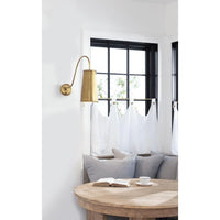 Hattie Sconce in natural brass with a cylinder shade, mounted on a white wall above a window nook.