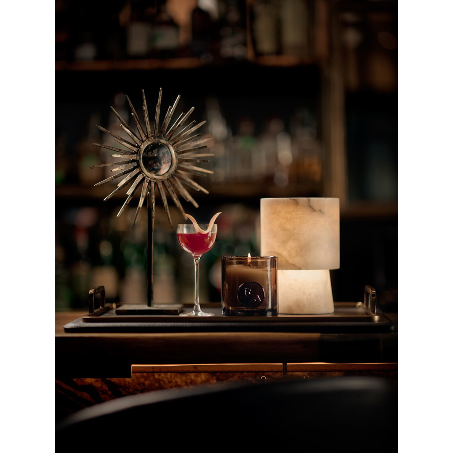 Hazel Alabaster Uplight Lamp, a translucent, soft stone lamp, on a dark tray with a cocktail and candle.