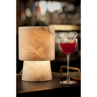Hazel Alabaster Uplight Lamp: translucent, cylindrical alabaster lamp with a warm glow, next to a red cocktail.