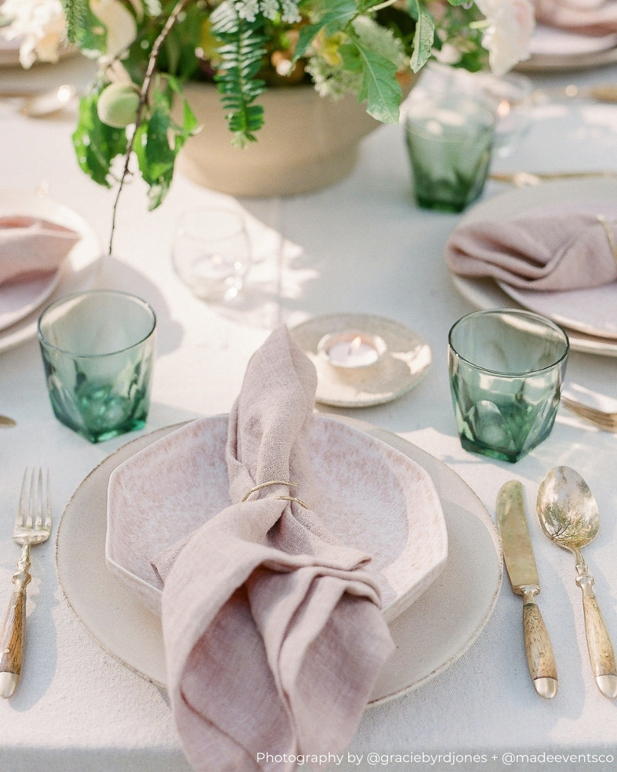Elegant table setting with green glasses, pink napkins, and silverware on a white tablecloth.