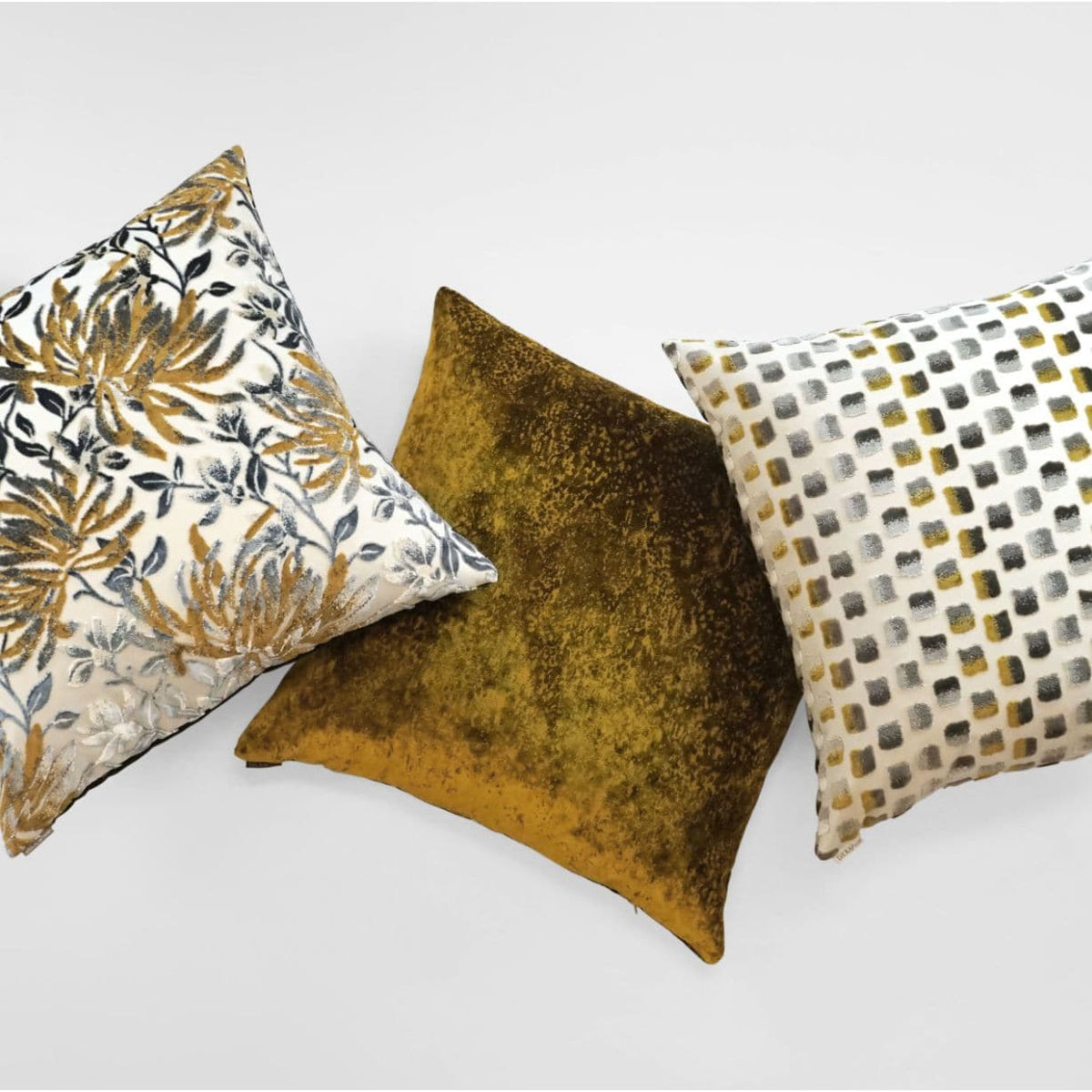 Three luxury designer throw pillows: a 24" mustard multi floral, a solid mustard velvet, and a white and gray geometric.