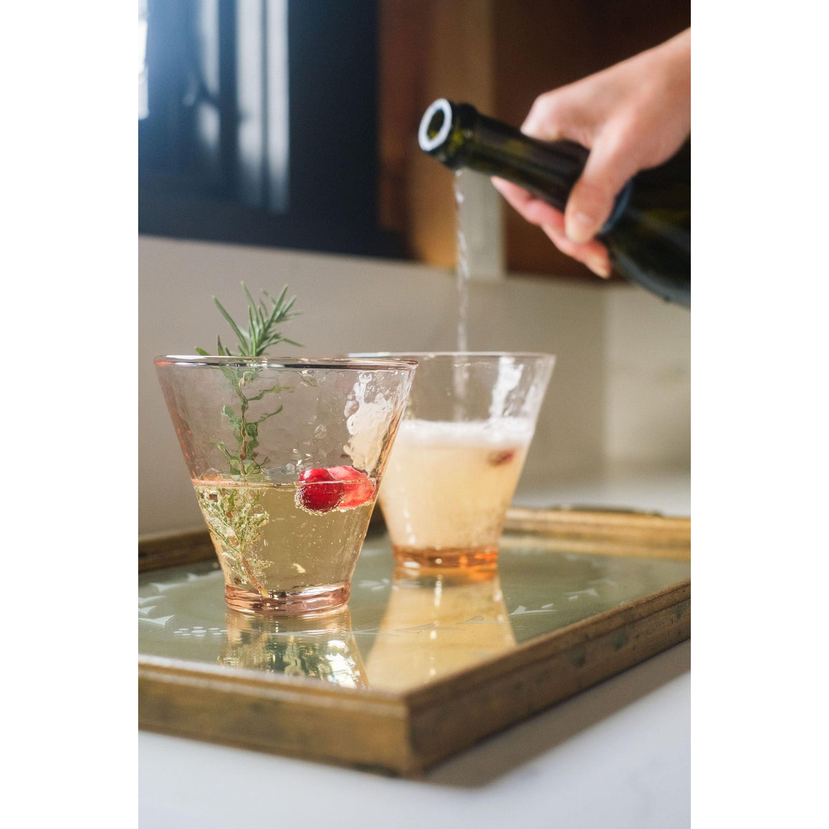 Person pouring a drink into a glass with a sprig of rosemary on a tray.