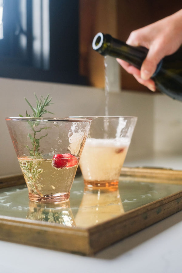 Person pouring a drink into a glass with a sprig of rosemary on a tray.