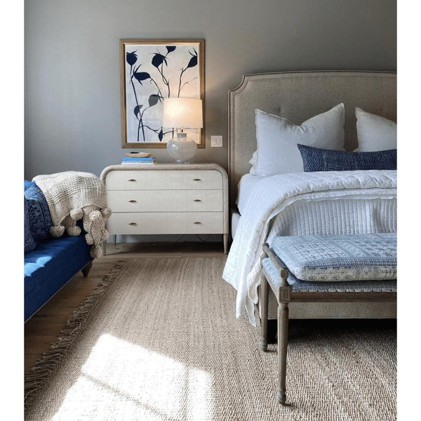 Beige hand-woven jute rug with fringe in a bedroom with a bed, bench, and dresser.
