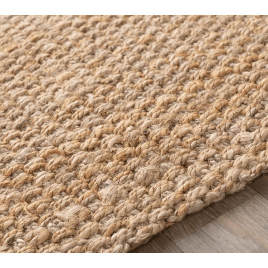 Close-up of a tan, hand-woven jute rug with a textured, no-pile design on a wooden floor.