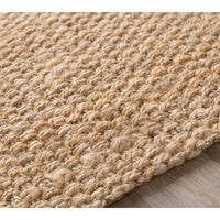 Close-up of a tan, hand-woven jute rug with a textured, no-pile design on a wooden floor.
