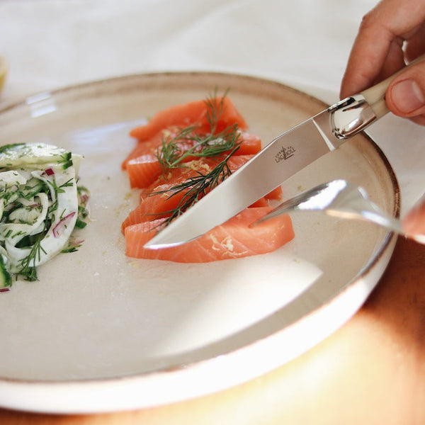 Sliced salmon with herbs on a plate, accompanied by a person using a knife and fork.