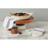 Wooden cake stand with a dish of food, blueberries, and oven mitts on a marble surface.