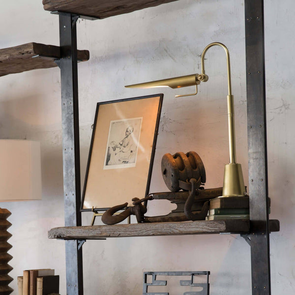Raven Task Lamp in natural brass with an adjustable shade, on a rustic wooden shelf.