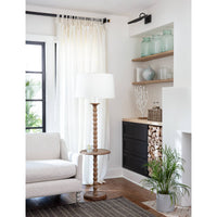 Tall wooden floor lamp with a white drum shade and a small round side table, next to a light-colored armchair.