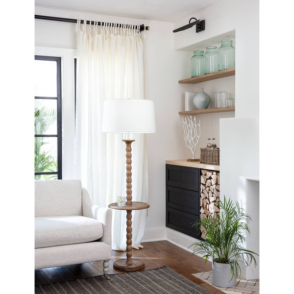 Tall wooden floor lamp with a white drum shade and a small round side table, next to a light-colored armchair.