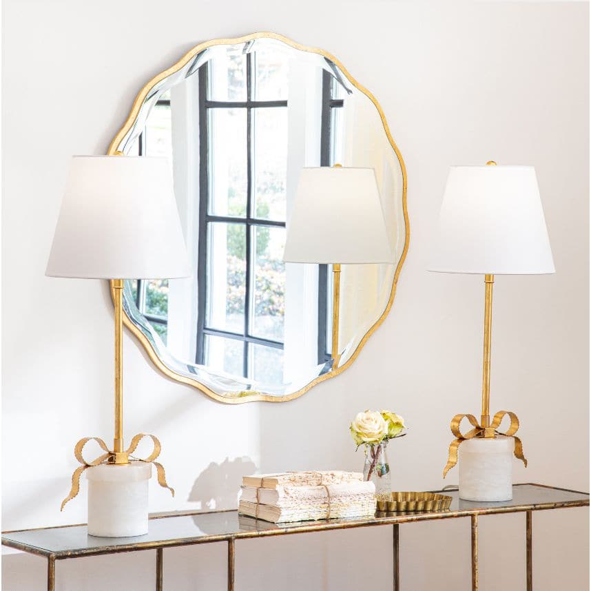 Elegant gold-leafed Ribbon Table Lamp with white linen shade and alabaster base, beside a scalloped mirror.