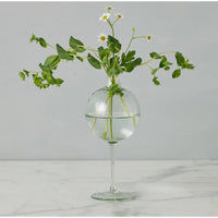 Clear glass wine glass vase with a spherical top, holding green foliage and white daisy-like flowers.