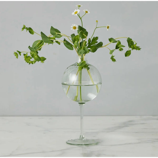 Clear glass wine glass vase with a spherical top, holding green foliage and white daisy-like flowers.