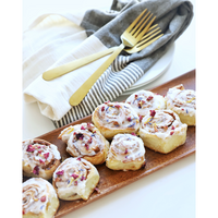 Wooden platter with cinnamon rolls topped with cream cheese frosting set on a white surface with a striped cloth and gold fork.