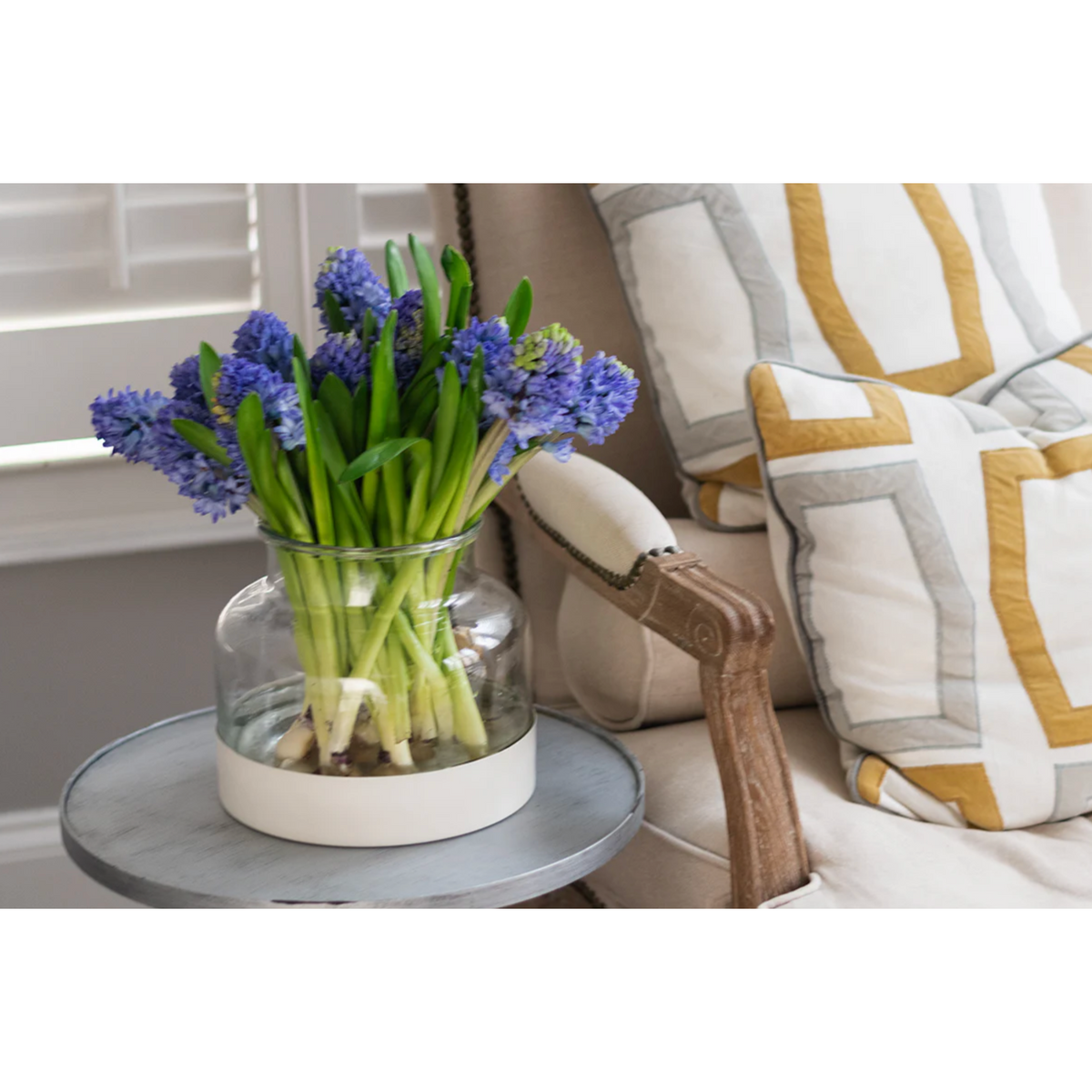 Clear vase with blue flowers on a small round table next to a striped armchair.
