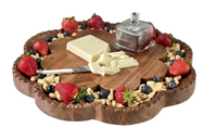 A scalloped wooden charcuterie board filled with peanuts, strawberries, blueberries, and cheese, with a small glass butter dish on a marble surface.