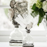 Tall clear glass bottle with a selenite crystal geode and slabs, next to white hydrangeas.