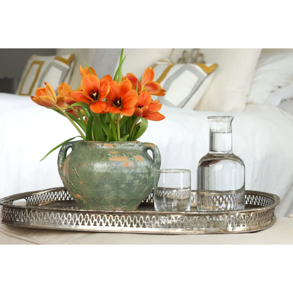 Antique green clay pot with handles, holding orange tulips, on a silver tray with a glass of water.