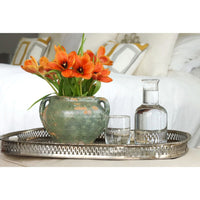 Antique green clay pot with handles, holding orange tulips, on a silver tray with a glass of water.