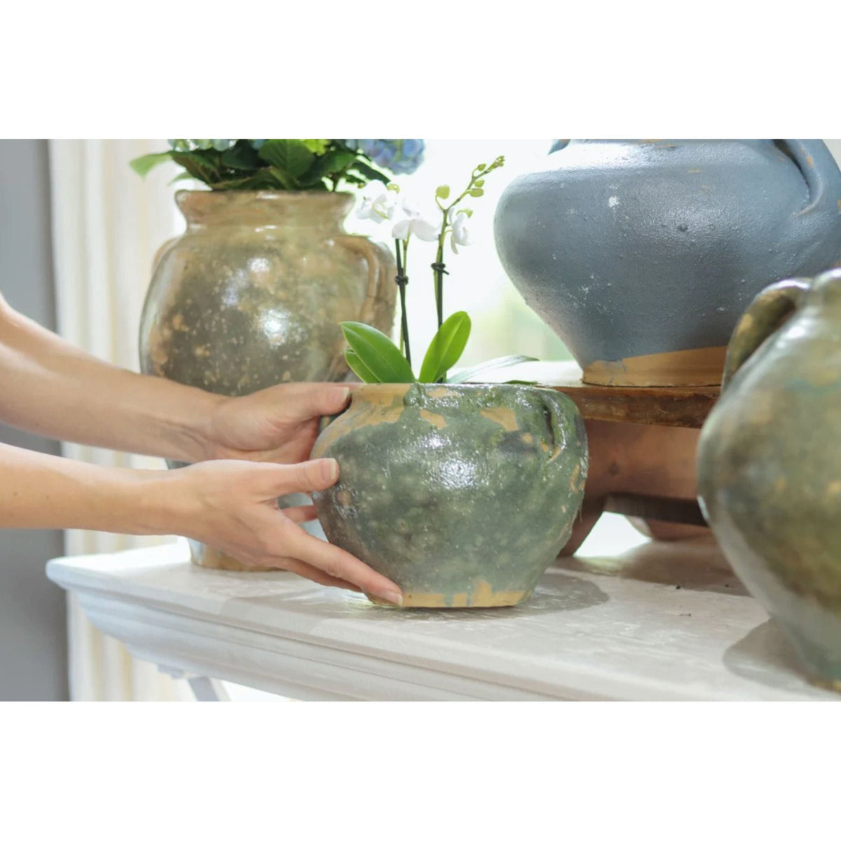Hands placing a small, round, antique green French clay pot with an orchid on a white mantel.