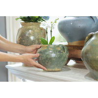 Hands placing a small, round, antique green French clay pot with an orchid on a white mantel.