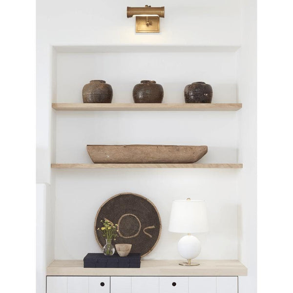 Small natural brass Tate Picture Light illuminating a white wall above shelves with decor.