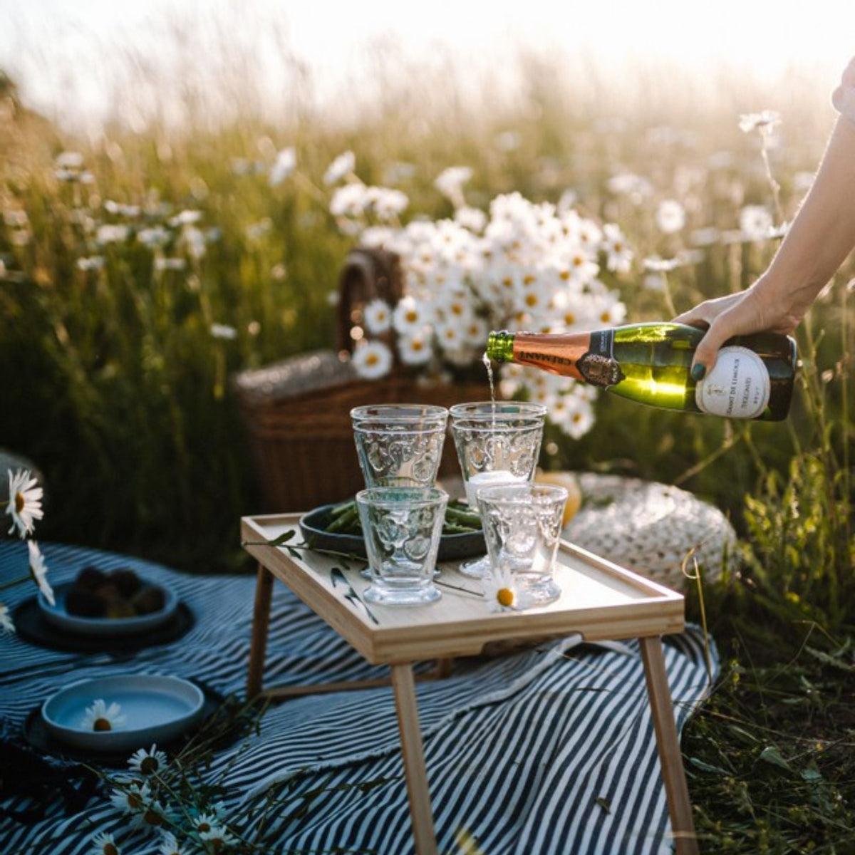 Person pouring wine into glasses on a small table in a field with flowers and picnic blanket.