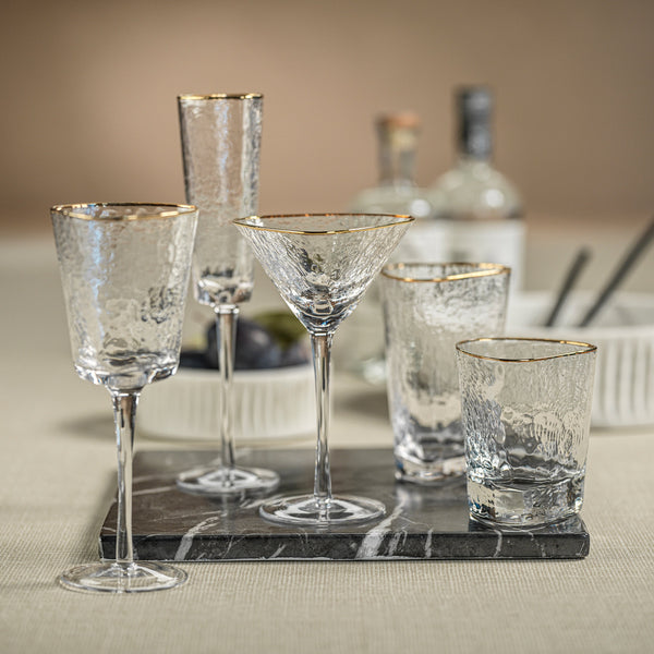 Set of clear glassware with gold rims including wine glasses, martini glasses, and tumblers on a marble tray.