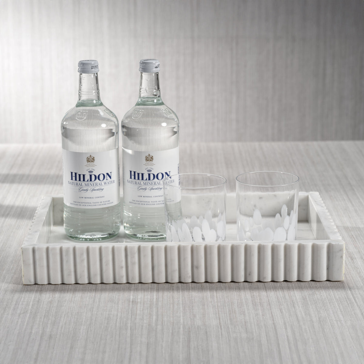 Two bottles of Hildon water on a marble tray with glasses against a neutral background